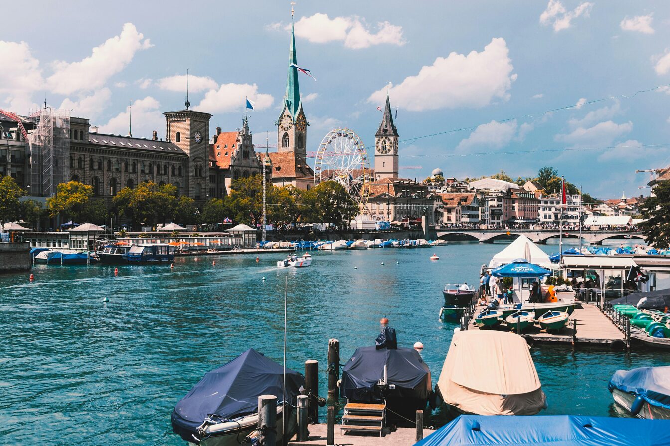 Blick auf den Fluss Limmat in Zürich mit überdachten Booten im Vordergrund. Im Hintergrund sind die Kirchtürme von Fraumünster und St. Peter zu sehen sowie ein großes Riesenrad. Der Himmel ist leicht bewölkt, und zahlreiche Menschen genießen den Tag entlang des Flussufers.