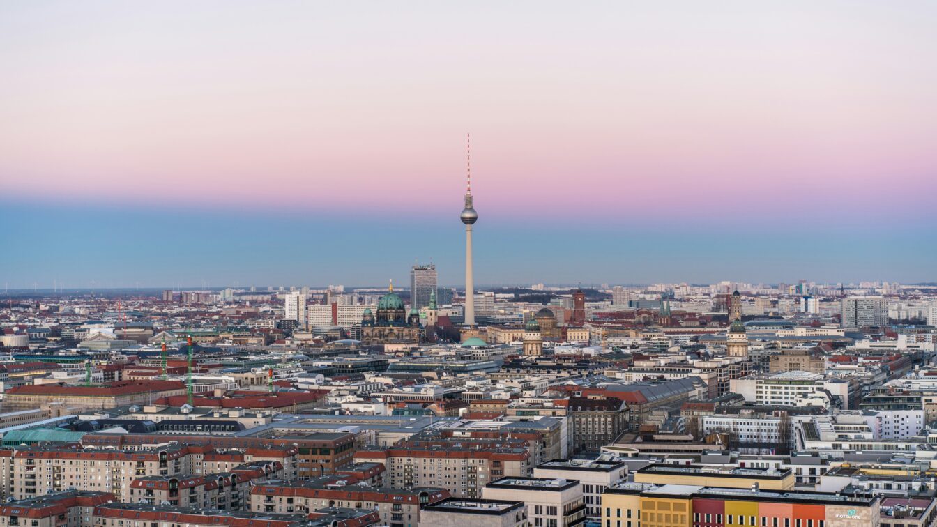 Das Bild zeigt die Skyline von Berlin bei Sonnenuntergang. Im Zentrum steht der Berliner Fernsehturm, umgeben von verschiedenen Gebäuden und Sehenswürdigkeiten der Stadt, wie dem Berliner Dom. Der Himmel hat einen sanften Farbverlauf von Rosa bis Hellblau, was dem Bild eine ruhige, stimmungsvolle Atmosphäre verleiht.