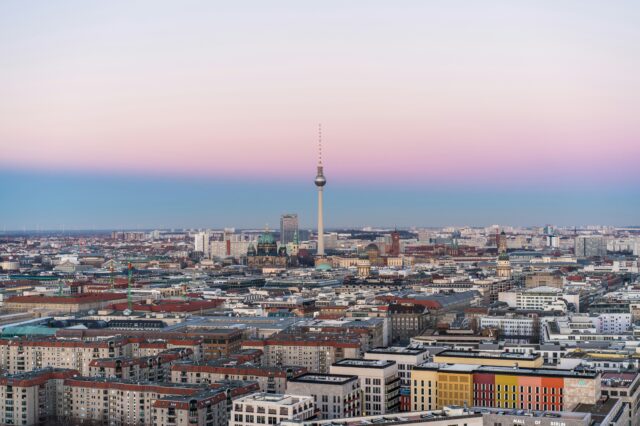 Das Bild zeigt die Skyline von Berlin bei Sonnenuntergang. Im Zentrum steht der Berliner Fernsehturm, umgeben von verschiedenen Gebäuden und Sehenswürdigkeiten der Stadt, wie dem Berliner Dom. Der Himmel hat einen sanften Farbverlauf von Rosa bis Hellblau, was dem Bild eine ruhige, stimmungsvolle Atmosphäre verleiht.