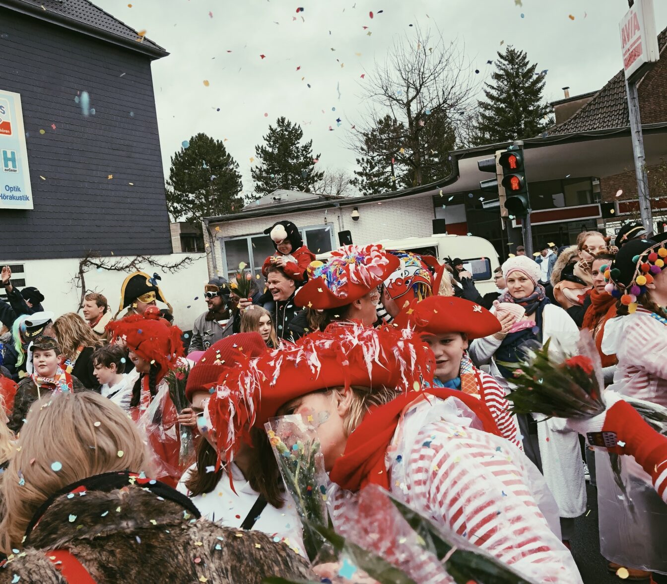 Fröhliche Menschen in bunten Kostümen feiern den Karneval in Köln, Deutschland. Sie tragen rot-weiße Kleidung und Hüte, halten Blumensträuße und lachen inmitten von Konfetti, das in der Luft schwebt. Die Menschenmenge steht vor Gebäuden und Ampeln auf der Straße, während die festliche Atmosphäre von Musik und Freude geprägt ist.