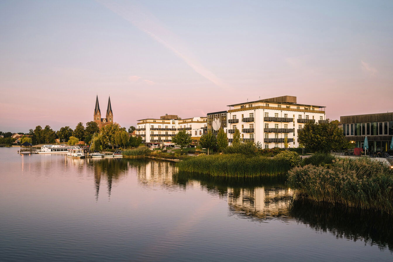 Blick auf das Resort Mark Brandenburg am Ufer eines ruhigen Sees in der Abenddämmerung. Die weißen Gebäude des Resorts reflektieren sich sanft im Wasser, umgeben von üppigem Grün und Schilf entlang der Küste. Im Hintergrund ragen die markanten Kirchtürme eines historischen Gebäudes empor. Der Himmel ist in sanfte Rosa- und Blautöne getaucht, was der Szene eine friedliche, romantische Atmosphäre verleiht.