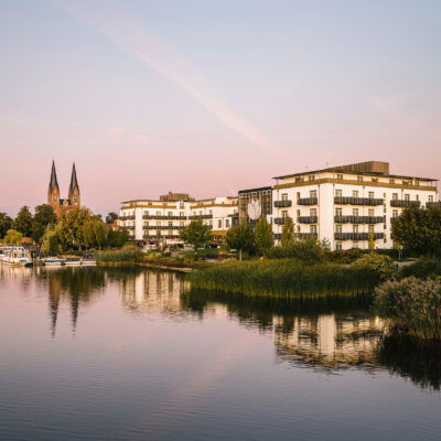 Blick auf das Resort Mark Brandenburg am Ufer eines ruhigen Sees in der Abenddämmerung. Die weißen Gebäude des Resorts reflektieren sich sanft im Wasser, umgeben von üppigem Grün und Schilf entlang der Küste. Im Hintergrund ragen die markanten Kirchtürme eines historischen Gebäudes empor. Der Himmel ist in sanfte Rosa- und Blautöne getaucht, was der Szene eine friedliche, romantische Atmosphäre verleiht.