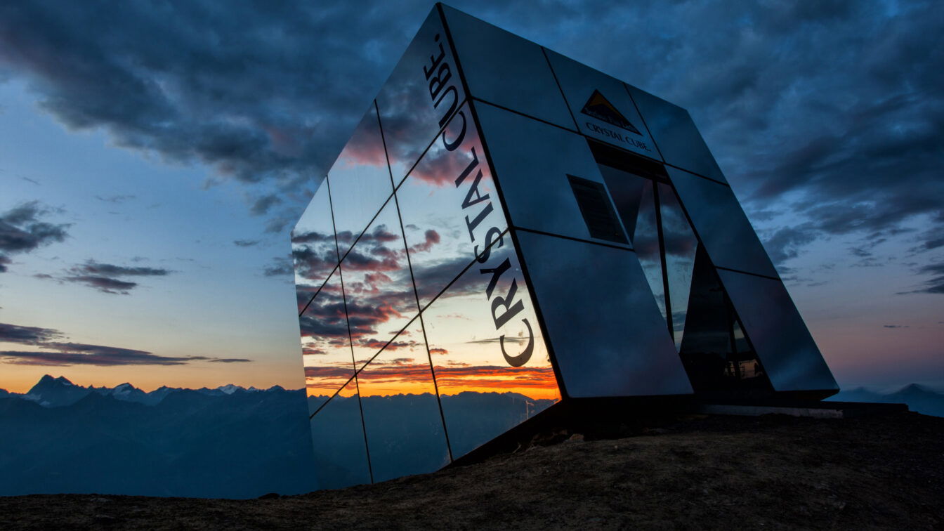 Das Bild zeigt den Crystal Cube in Serfaus-Fiss-Ladis bei Sonnenuntergang. Der kubusförmige, verspiegelte Bau steht auf einem Berggipfel und reflektiert das atemberaubende Panorama der umliegenden Alpen. Der Himmel ist von dramatischen Wolken bedeckt, die sich in den Spiegelwänden des Crystal Cube widerspiegeln. Die warmen Farben des Sonnenuntergangs kontrastieren mit den kühlen Tönen des Himmels und verleihen der Szene eine mystische Atmosphäre.