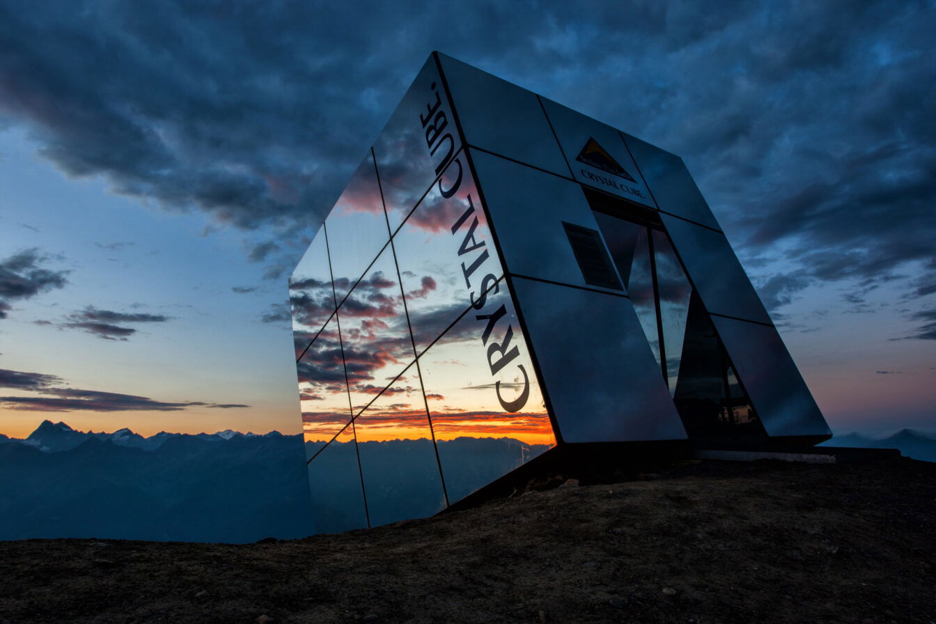 Das Bild zeigt den Crystal Cube in Serfaus-Fiss-Ladis bei Sonnenuntergang. Der kubusförmige, verspiegelte Bau steht auf einem Berggipfel und reflektiert das atemberaubende Panorama der umliegenden Alpen. Der Himmel ist von dramatischen Wolken bedeckt, die sich in den Spiegelwänden des Crystal Cube widerspiegeln. Die warmen Farben des Sonnenuntergangs kontrastieren mit den kühlen Tönen des Himmels und verleihen der Szene eine mystische Atmosphäre.