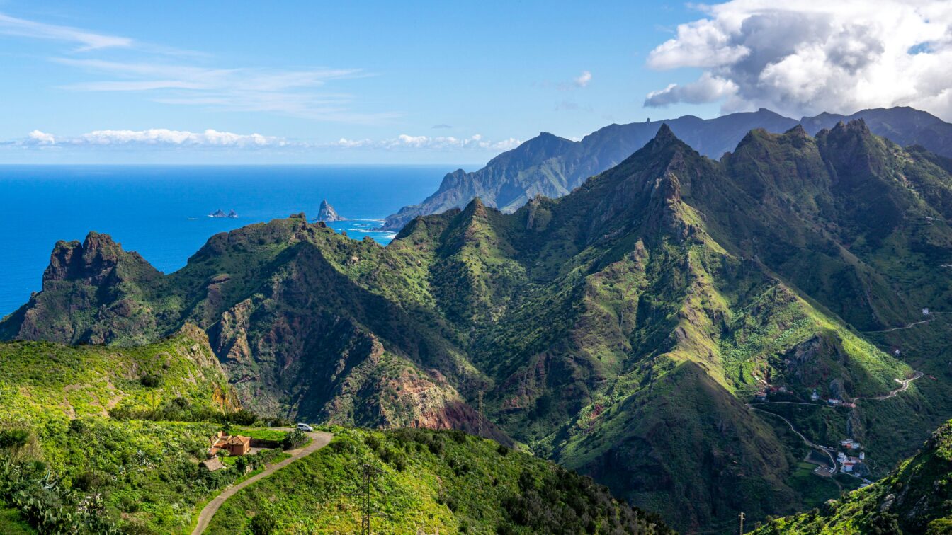 Panorama der grünen, zerklüfteten Berglandschaft auf Teneriffa, Kanarische Inseln. Im Vordergrund eine kleine Straße und ein Haus, während die bewachsenen Berge sich zum Meer hin erstrecken. Am Horizont liegt der blaue Atlantik, gesäumt von Felsen im Wasser und sanften Wolken am Himmel.