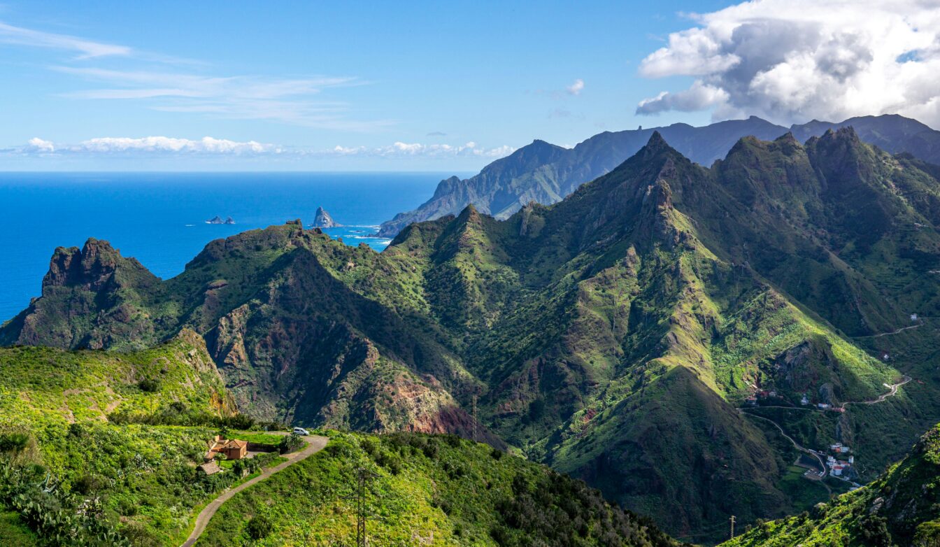 Panorama der grünen, zerklüfteten Berglandschaft auf Teneriffa, Kanarische Inseln. Im Vordergrund eine kleine Straße und ein Haus, während die bewachsenen Berge sich zum Meer hin erstrecken. Am Horizont liegt der blaue Atlantik, gesäumt von Felsen im Wasser und sanften Wolken am Himmel.