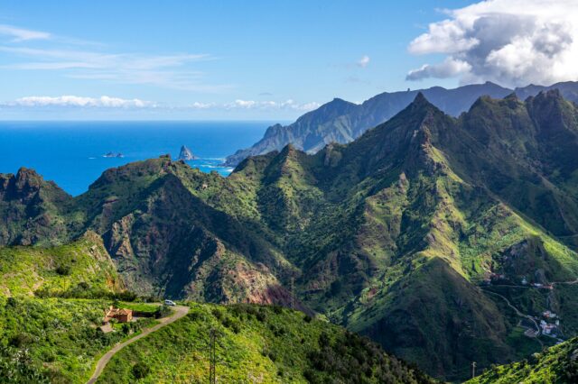 Panorama der grünen, zerklüfteten Berglandschaft auf Teneriffa, Kanarische Inseln. Im Vordergrund eine kleine Straße und ein Haus, während die bewachsenen Berge sich zum Meer hin erstrecken. Am Horizont liegt der blaue Atlantik, gesäumt von Felsen im Wasser und sanften Wolken am Himmel.