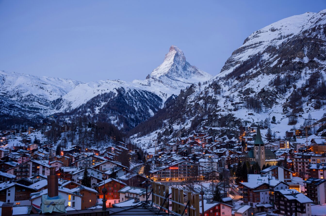 Das Bild zeigt die winterliche Landschaft von Zermatt in der Schweiz bei Dämmerung. Im Hintergrund erhebt sich majestätisch das Matterhorn, dessen Gipfel von einer leichten Schneeschicht bedeckt ist. Die Stadt Zermatt breitet sich im Tal aus, ihre Dächer sind ebenfalls mit Schnee bedeckt und warme Lichter leuchten aus den Fenstern der Häuser und Hotels, was eine gemütliche Atmosphäre schafft. Im Vordergrund ist die Kirche von Zermatt mit ihrem markanten Turm sichtbar.