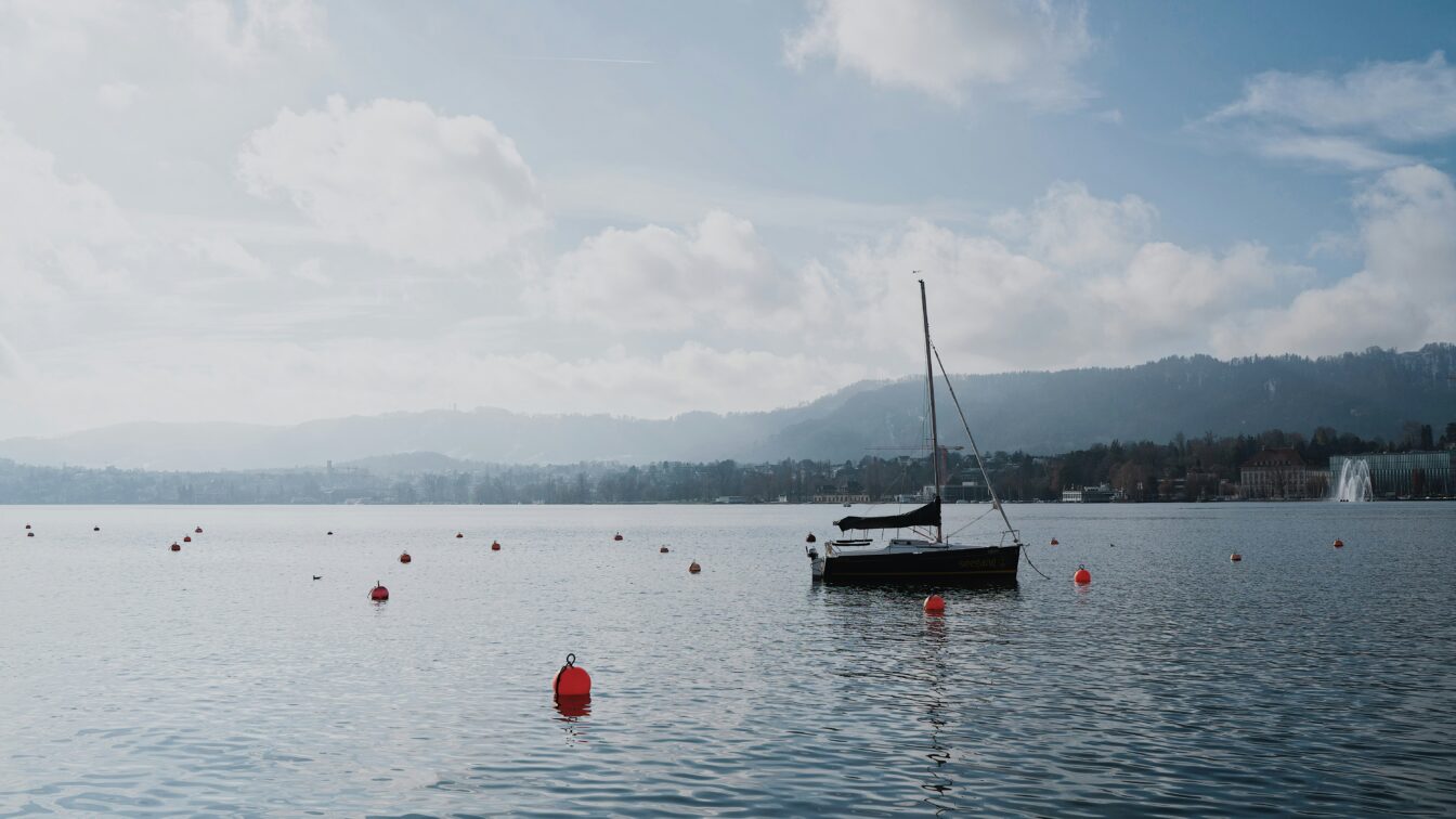 Ein ruhiger See mit einer Segelyacht und mehreren roten Bojen im Wasser. Der Himmel ist leicht bewölkt, und die umliegenden Hügel und Gebäude bilden eine sanfte Kulisse im Hintergrund. Auf der rechten Seite sprüht eine Wasserfontäne. Die Atmosphäre wirkt friedlich und klar, mit weichen Lichtverhältnissen und ruhigem Wasser, das die Umgebung sanft reflektiert.