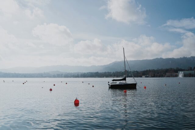 Ein ruhiger See mit einer Segelyacht und mehreren roten Bojen im Wasser. Der Himmel ist leicht bewölkt, und die umliegenden Hügel und Gebäude bilden eine sanfte Kulisse im Hintergrund. Auf der rechten Seite sprüht eine Wasserfontäne. Die Atmosphäre wirkt friedlich und klar, mit weichen Lichtverhältnissen und ruhigem Wasser, das die Umgebung sanft reflektiert.