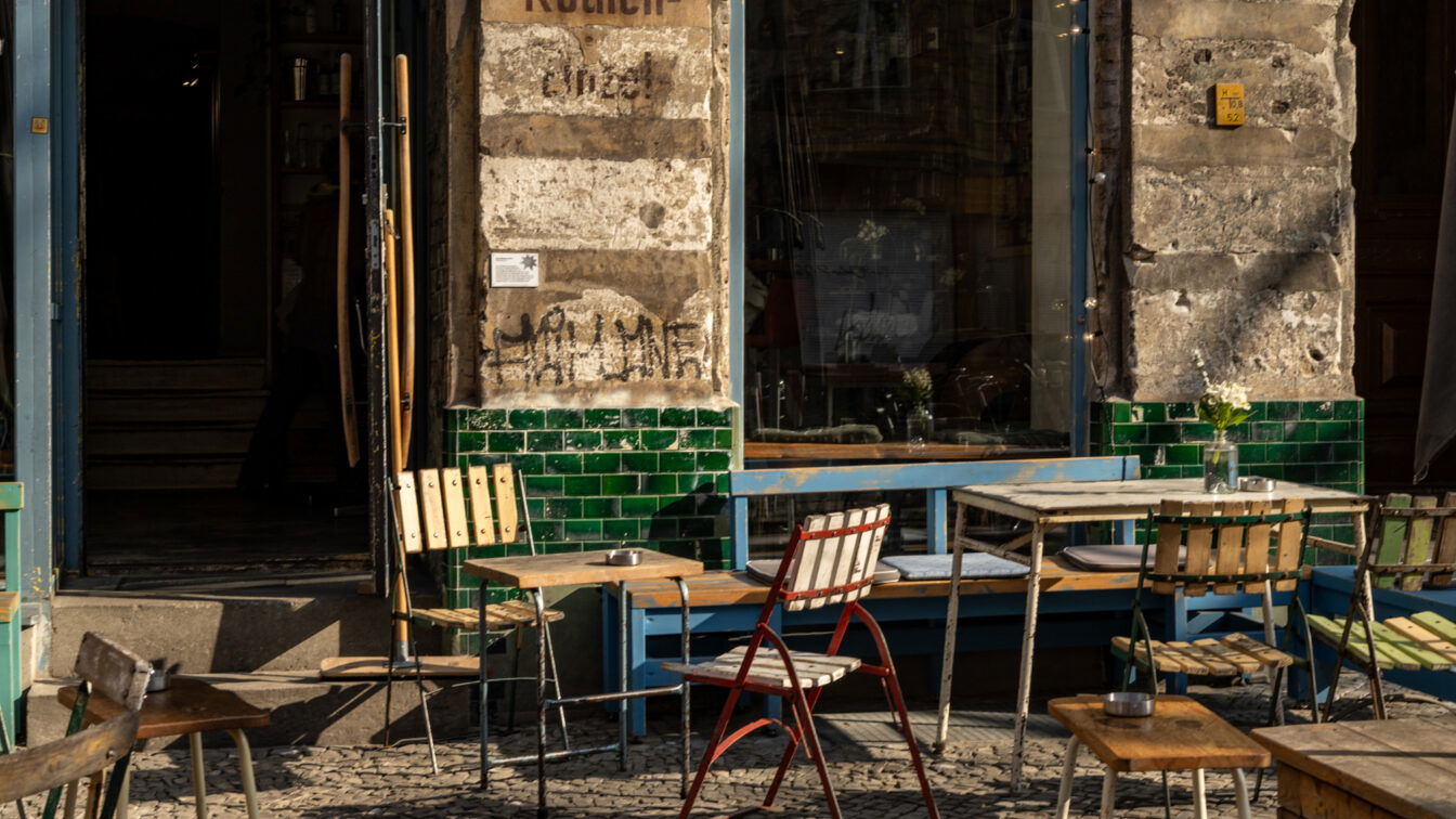 (c) Sonja Koller | 1000things Ein charmantes Berliner Café mit einer rustikalen Fassade aus verwittertem Putz und grünen Fliesen. Vor dem Eingang stehen mehrere alte, teils wackelige Holztische und Stühle, darunter ein auffälliger weiß-roter Klappstuhl. Die Fenster spiegeln die Umgebung wider, und auf den Tischen stehen kleine Vasen mit weißen Blumen. Die Sonne wirft warme Lichtstrahlen und erzeugt lange Schatten auf dem Kopfsteinpflaster, während im Hintergrund eine geöffnete Tür den Blick ins Innere des Cafés freigibt.