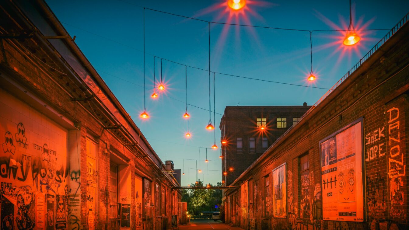 Eine schmale Gasse zwischen alten Backsteingebäuden mit Graffiti, beleuchtet von orangefarbenen Lampen, die stimmungsvoll im Abendlicht hängen.