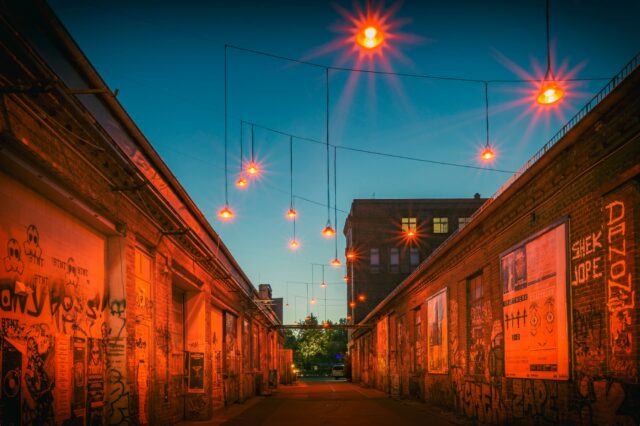 Eine schmale Gasse zwischen alten Backsteingebäuden mit Graffiti, beleuchtet von orangefarbenen Lampen, die stimmungsvoll im Abendlicht hängen.