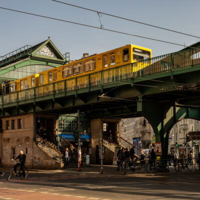 Die U-Bahn-Station Eberswalder Straße in Berlin mit einer gelben U-Bahn, die über eine grüne Stahlbrücke fährt. Die historische Hochbahnstation hat eine markante Architektur mit verzierten Fenstern und einem Giebeldach mit Graffiti. Unter der Brücke beleben Fußgänger, Radfahrer und Autos die Kreuzung. Mehrere Menschen steigen die Treppen zur Station hinauf oder verlassen sie. Im Hintergrund sind Geschäfte, Cafés und Werbetafeln zu sehen, während die tief stehende Sonne lange Schatten auf die Szene wirft