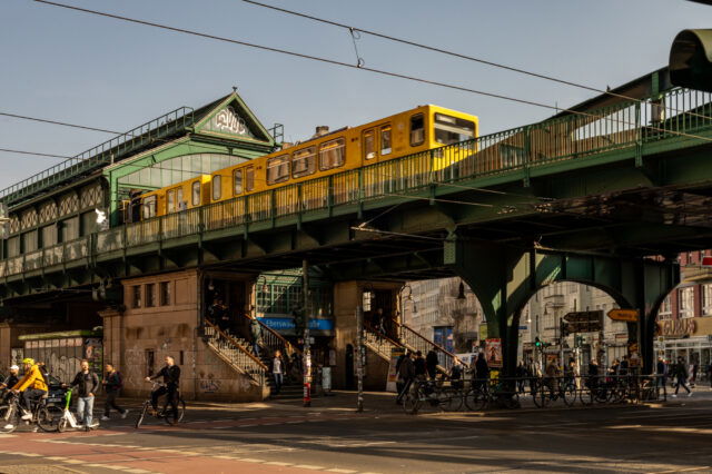 Die U-Bahn-Station Eberswalder Straße in Berlin mit einer gelben U-Bahn, die über eine grüne Stahlbrücke fährt. Die historische Hochbahnstation hat eine markante Architektur mit verzierten Fenstern und einem Giebeldach mit Graffiti. Unter der Brücke beleben Fußgänger, Radfahrer und Autos die Kreuzung. Mehrere Menschen steigen die Treppen zur Station hinauf oder verlassen sie. Im Hintergrund sind Geschäfte, Cafés und Werbetafeln zu sehen, während die tief stehende Sonne lange Schatten auf die Szene wirft