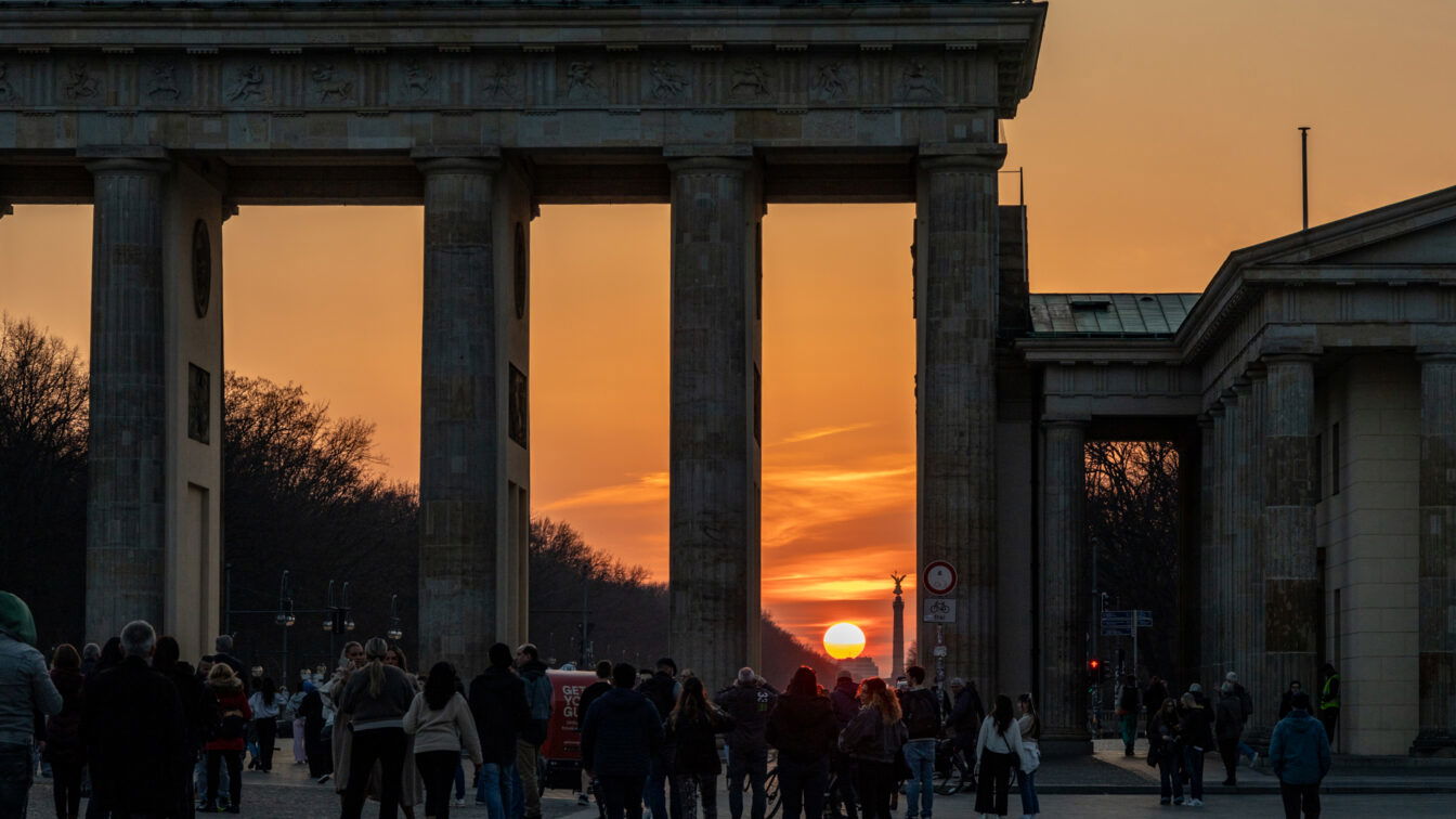 (c) Sonja Koller | 1000things Menschen spazieren bei Sonnenuntergang durch das Brandenburger Tor in Berlin. Die Sonne steht tief am Horizont und scheint genau durch die Säulen des Tores. Der Himmel ist orange gefärbt, Silhouetten der Menschen und Gebäude rahmen die Szene stimmungsvoll ein.