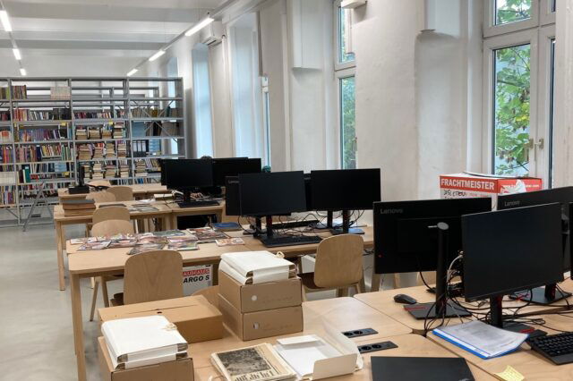 Das Bild zeigt einen modernen Raum, der wie ein Arbeitsbereich in einer Bibliothek oder einem Archiv aussieht. Es gibt mehrere Tische mit Computern und Büchern. Auf den Tischen liegen Zeitschriften und einige Dokumente, die teilweise in weiße Ordner verpackt sind. Im Hintergrund sind Regale mit einer Vielzahl von Büchern sichtbar. Große Fenster lassen natürliches Licht in den Raum, und im hinteren Bereich des Raums ist ein Kasten mit Aufschrift "Frachtmeister" zu sehen. Ein Mitarbeiter ist an einem der Computer zu sehen, während er an einem Bildschirm arbeitet.