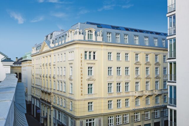 Außenansicht des Steigenberger Hotels, ein elegantes, hellgelbes Gebäude mit klassischer Architektur, zahlreichen Fenstern und kunstvollen Fassadendetails. Der Hotelname ist vertikal an der Seite der Fassade zu sehen, der Himmel ist blau und klar.