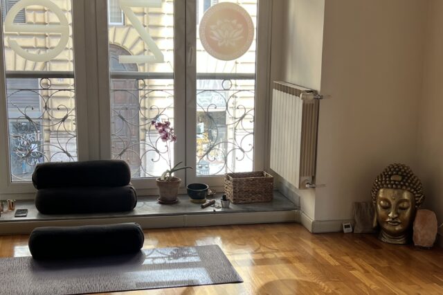 Dieses Bild zeigt einen einladenden Yoga- oder Meditationsraum mit einer warmen und beruhigenden Atmosphäre. Im Vordergrund liegt eine Yogamatte mit einem schwarzen Bolster darauf. Weitere Bolster sind auf der Fensterbank gestapelt. Auf der Fensterbank befinden sich zudem eine Orchidee, eine kleine Schale und ein geflochtener Korb mit verschiedenen Gegenständen. Rechts im Raum steht eine große goldene Buddha-Kopf-Skulptur neben einem Salzkristalllicht. Durch die großen Fenster mit kunstvollen Gitterverzierungen fällt natürliches Licht ein, und im Hintergrund ist eine städtische Umgebung mit verzierten Gebäudefassaden zu sehen.