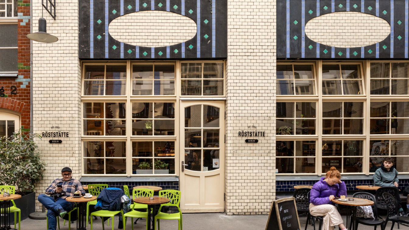 Außenansicht des Cafés „Röststätte Berlin“ in einem charmanten Hinterhof mit heller Klinkerfassade und auffälligen Verzierungen. Vor dem Café sitzen mehrere Menschen an kleinen Tischen, genießen Kaffee und wirken entspannt. Eine Tafel bewirbt Espresso. Die Atmosphäre ist ruhig und urban.
