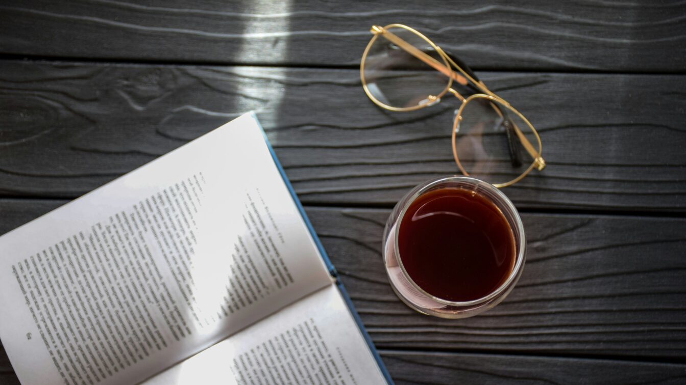 (c) Catia Climovich | 1000things Ein aufgeschlagenes Buch neben einem Glas Rotwein und einer Lesebrille.