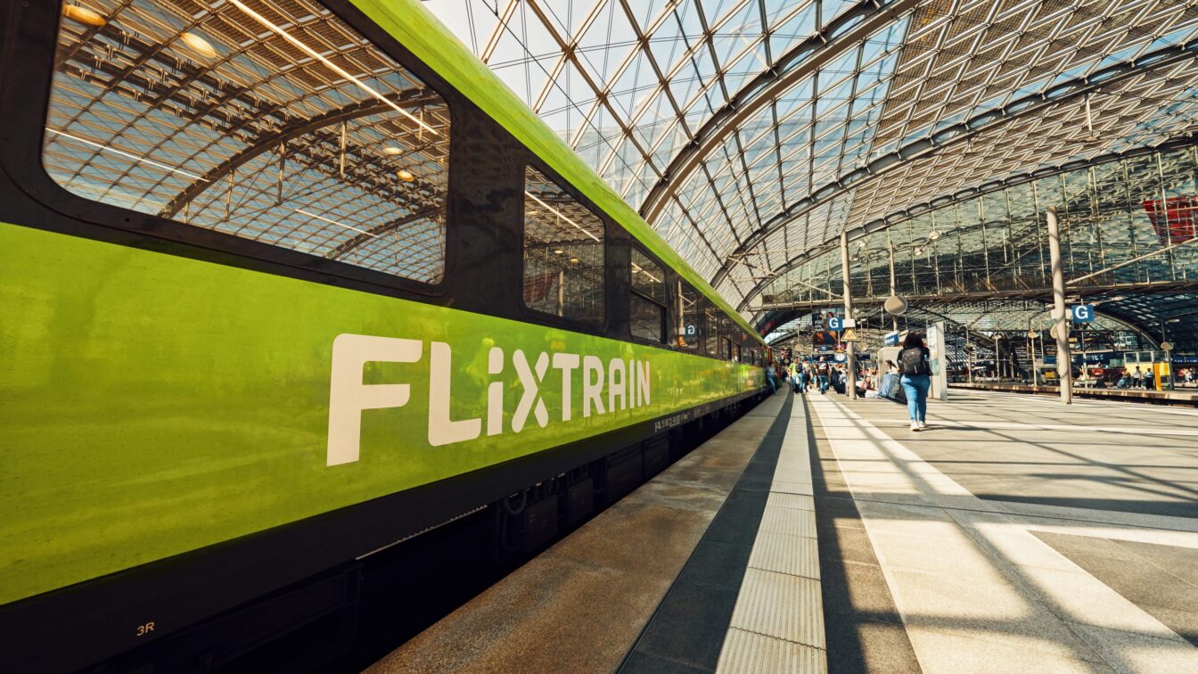 (c) Flixbus Ein grüner FlixTrain steht an einem Bahnsteig in einem modernen, lichtdurchfluteten Bahnhof mit gläsernem Dach. Menschen laufen entlang des Bahnsteigs, einige mit Gepäck. Die Perspektive zeigt den Zug in Nahaufnahme auf der linken Seite, während sich das Glasdach bogenförmig über die gesamte Szene spannt.