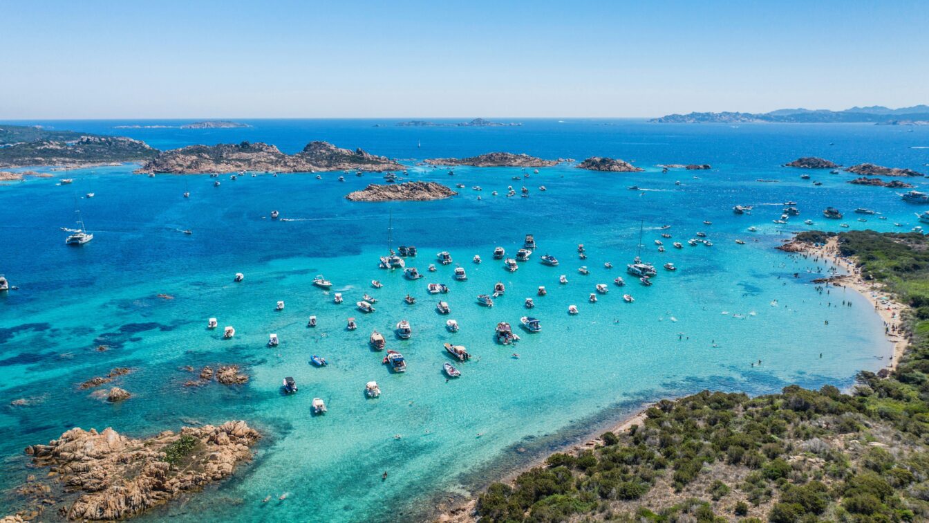(c) Ivan Ragozin | Unsplash Luftaufnahme vom türkisblauen Meer vor Sardinien mit zahlreichen Booten vor der Küste und feinem Sandstrand – beliebtes Sommerziel in Italien mit kristallklarem Wasser und mediterraner Landschaft.