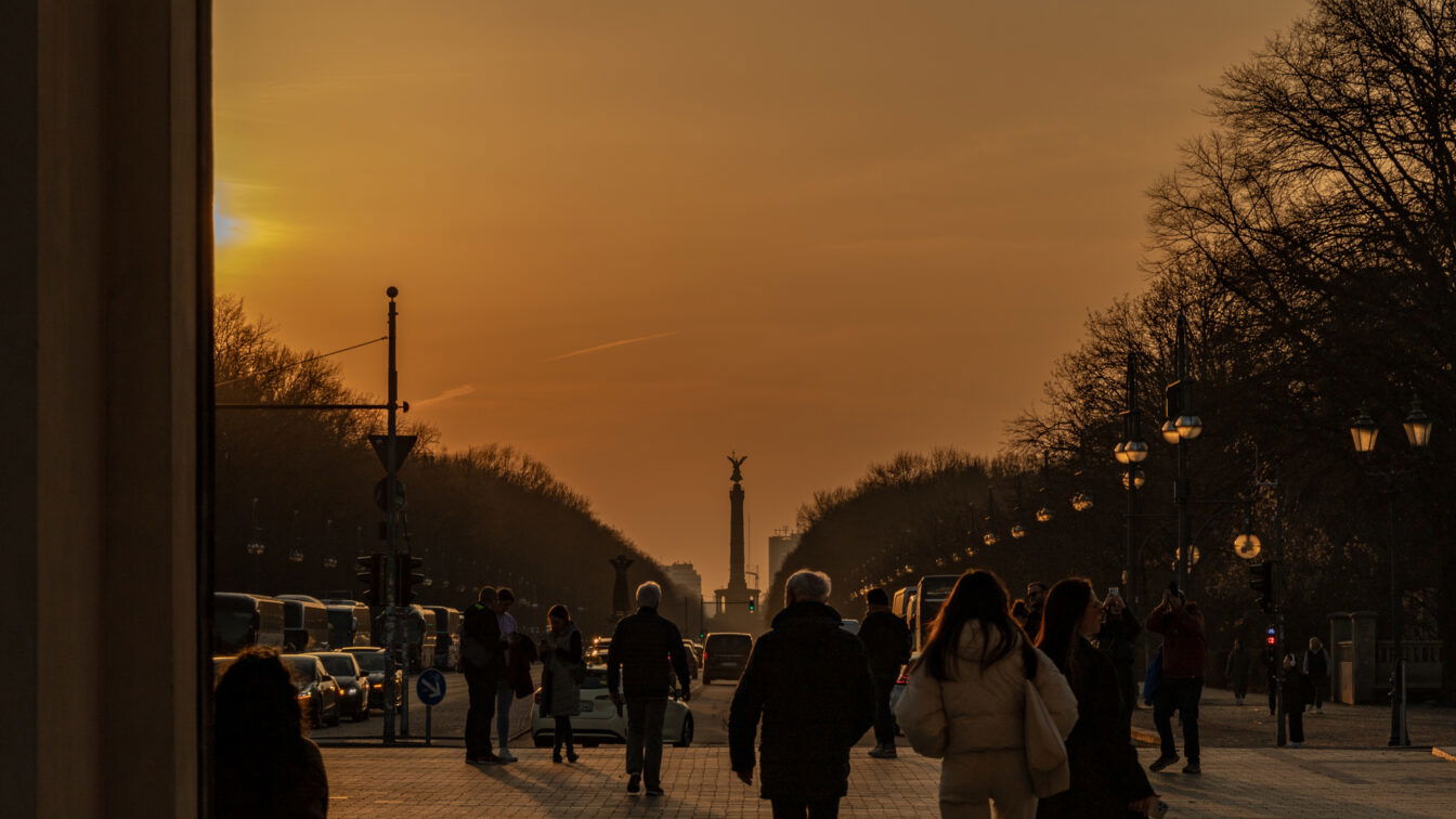 (c) Sonja Koller | 1000things Menschen spazieren bei Sonnenuntergang auf der Straße des 17. Juni in Berlin, mit Blick auf die goldene Siegessäule in der Ferne. Die tiefstehende Sonne taucht den Himmel in warme Orangetöne, während Laternenlichter entlang der Straße leuchten. Bäume säumen den Weg, Silhouetten und Schatten betonen die ruhige Abendstimmung.
