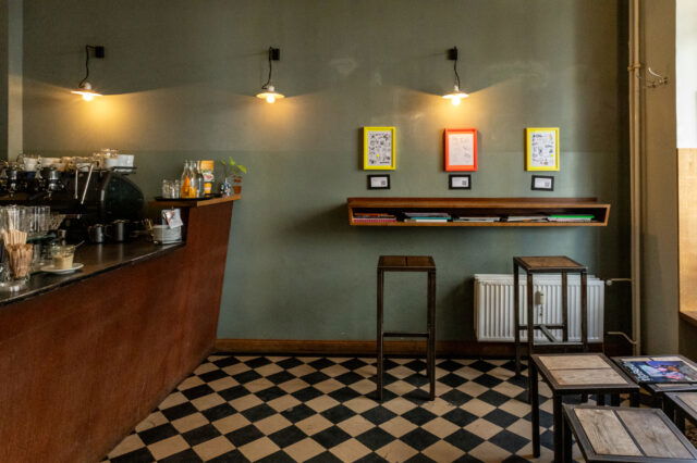 (c) Sonja Koller Gemütliches Café mit dunklem Thekenbereich links, Espressomaschine, Gläsern und Getränken. Die Wand ist in einem matten Grünton gestrichen und mit drei farbigen Bilderrahmen (gelb, rot, gelb) dekoriert, darunter kleine QR-Code-Schilder. Darunter ein schräges Wandregal mit Magazinen und Büchern. Zwei schlichte Barhocker stehen davor. Schwarz-weiß karierter Boden, sanfte Beleuchtung durch drei Wandleuchten. Rechts im Bild einfache Holztische mit Magazinen. Ruhige, minimalistische Atmosphäre.