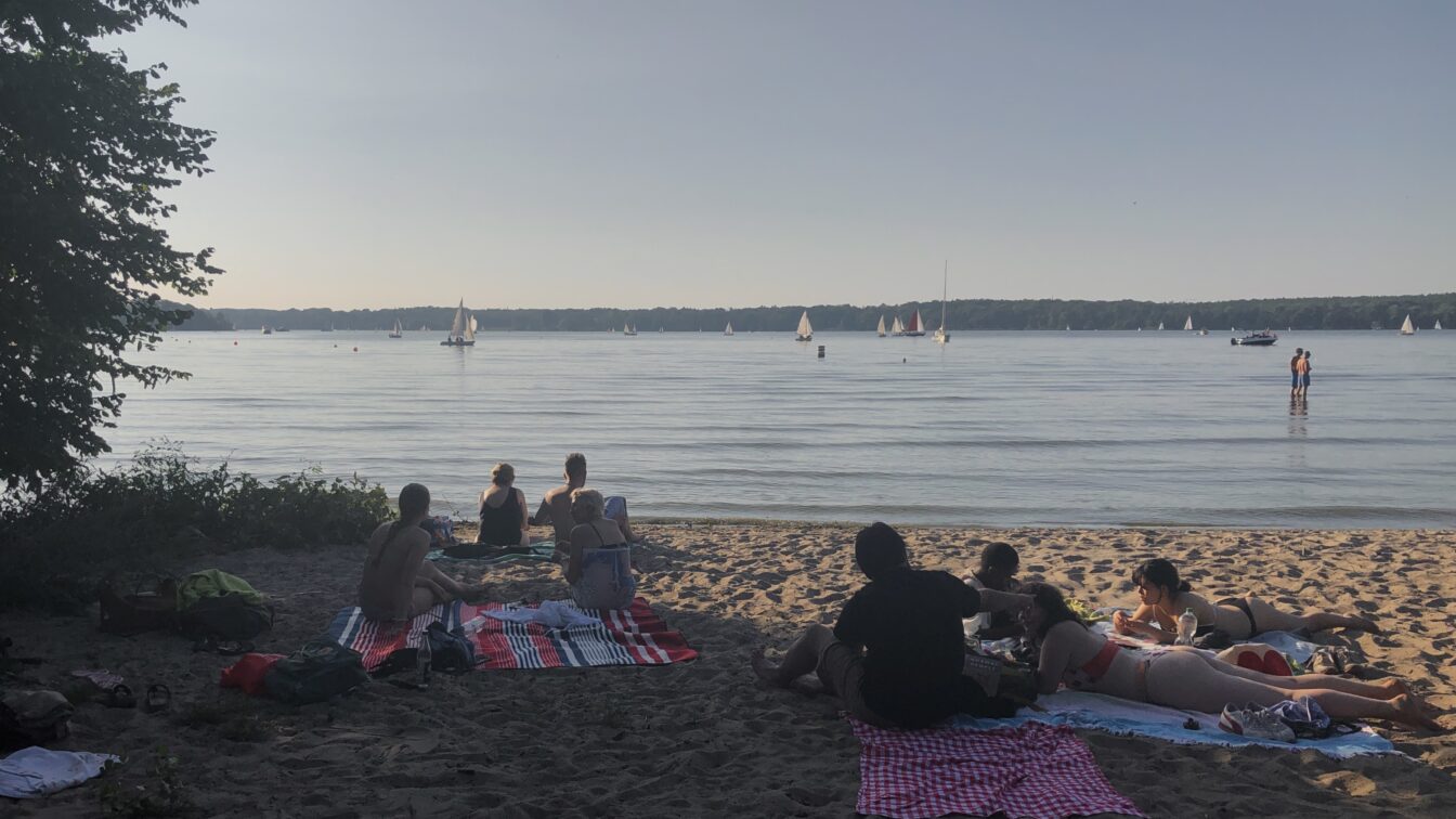 (c) Sonja Koller | 1000things Menschen sitzen und liegen entspannt auf Picknickdecken am Sandstrand eines ruhigen Sees, während im Hintergrund zahlreiche Segelboote auf dem Wasser treiben. Die Szene spielt sich an einem sonnigen, warmen Tag ab, das Licht ist weich und sommerlich. Einige Personen stehen im flachen Wasser, andere genießen die Aussicht aufs Wasser. Links ragen Bäume ins Bild und spenden Schatten.