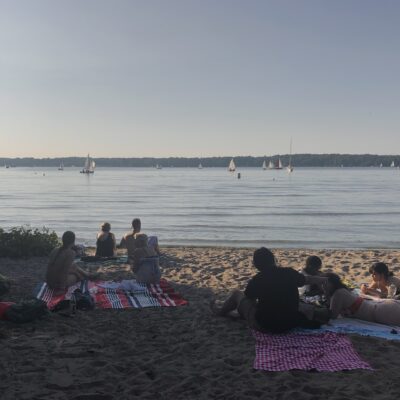 Menschen sitzen und liegen entspannt auf Picknickdecken am Sandstrand eines ruhigen Sees, während im Hintergrund zahlreiche Segelboote auf dem Wasser treiben. Die Szene spielt sich an einem sonnigen, warmen Tag ab, das Licht ist weich und sommerlich. Einige Personen stehen im flachen Wasser, andere genießen die Aussicht aufs Wasser. Links ragen Bäume ins Bild und spenden Schatten.