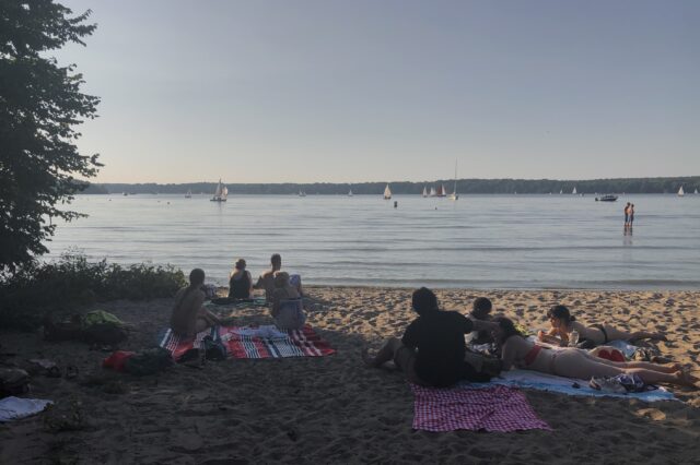 Menschen sitzen und liegen entspannt auf Picknickdecken am Sandstrand eines ruhigen Sees, während im Hintergrund zahlreiche Segelboote auf dem Wasser treiben. Die Szene spielt sich an einem sonnigen, warmen Tag ab, das Licht ist weich und sommerlich. Einige Personen stehen im flachen Wasser, andere genießen die Aussicht aufs Wasser. Links ragen Bäume ins Bild und spenden Schatten.