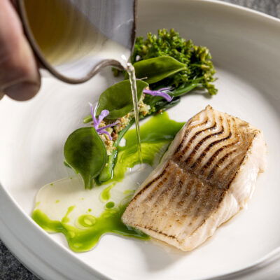 Gourmetgericht mit zart gebratener Forelle, grüner Kräutersauce, frischen Wildkräutern und essbaren Blüten – edle Fine-Dining-Komposition mit Sauce am Teller angerichtet, serviert auf handgefertigtem Porzellan.