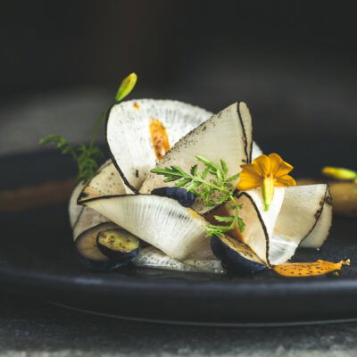 Gourmetgericht im Restaurant Kräuterreich by Vitus Winkler: kunstvoll angerichtete Komposition aus hauchdünnem Gemüse, Wildkräutern, Blüten und feinen Texturen auf dunklem Teller – moderne Alpenküche auf Sterne-Niveau.