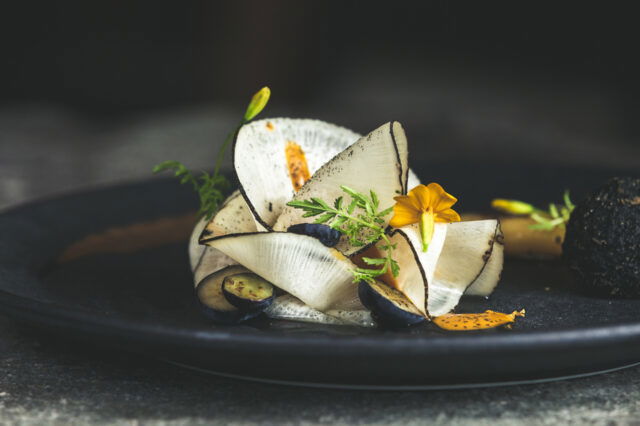 (c) Mario Stockhausen Gourmetgericht im Restaurant Kräuterreich by Vitus Winkler: kunstvoll angerichtete Komposition aus hauchdünnem Gemüse, Wildkräutern, Blüten und feinen Texturen auf dunklem Teller – moderne Alpenküche auf Sterne-Niveau.