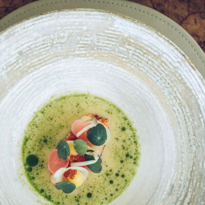 Kunstvoll angerichtetes Fine-Dining-Gericht im Restaurant The Glass Garden: zarte Komposition aus Gemüseperlen, Kräuteröl, Kaviar und Blüten auf cremiger Sauce – serviert in elegantem Keramikteller auf edlem Naturleder-Untersetzer.